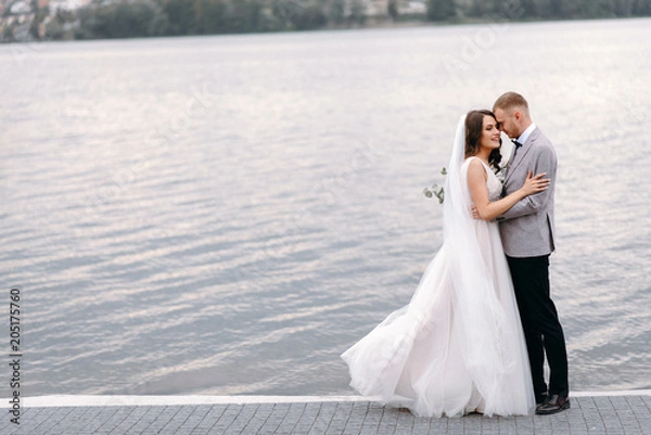 Obraz An amazing couple on their wedding day near the lake hug and enjoy each other. The bride and groom with a bouquet are happy together