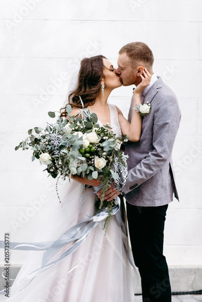 Obraz Portrait of a beautiful couple in love on your wedding day. Walking near the house with white walls and greenery. Amazing kisses and embraces of the bride and groom with a bouquet