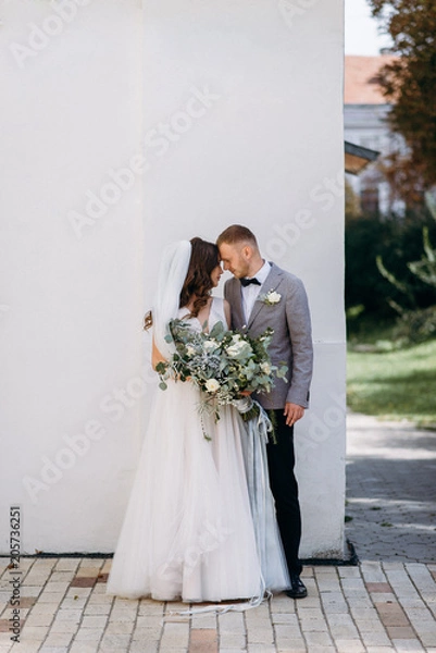 Obraz Portrait of a beautiful couple in love on your wedding day. Walking near the house with white walls and greenery. Amazing kisses and embraces of the bride and groom with a bouquet