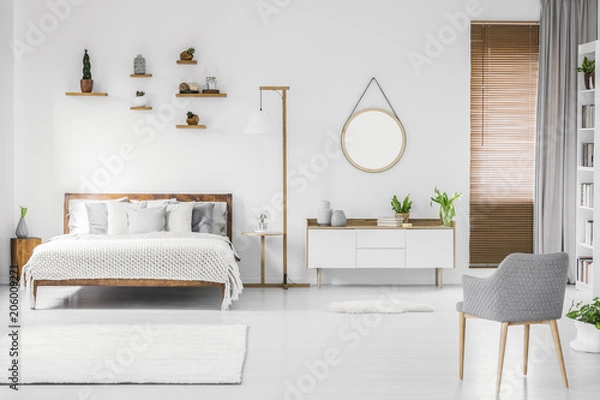 Fototapeta Przestronne, designerskie białe wnętrze sypialni z drewnianym łóżkiem z pościelą i poduszkami, stolikiem nocnym, półkami powyżej i nowoczesnym fotelem po prawej stronie. Prawdziwe zdjęcie.