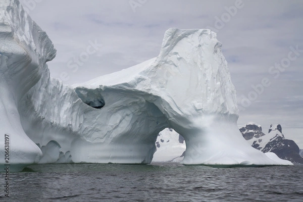 Obraz iceberg with an arch in Antarctic waters against the backdrop of the mountains of the Antarctic Peninsula on a cloudy day