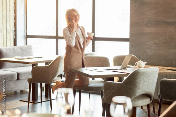 Fototapeta Young elegant businesswoman with cup of tea or coffee standing by table in cafe, talking by smartphone and making new appointment with one of clients