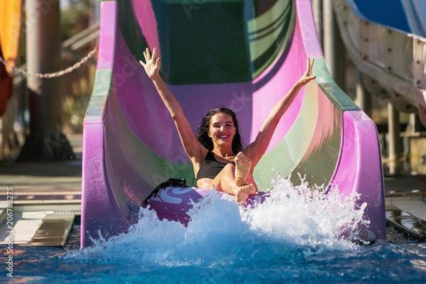 Obraz pretty brunette slim woman with raised hands on the rubber ring having fun coming down on the purple water slide in the aqua park. Summer Vacation. Weekend on resort