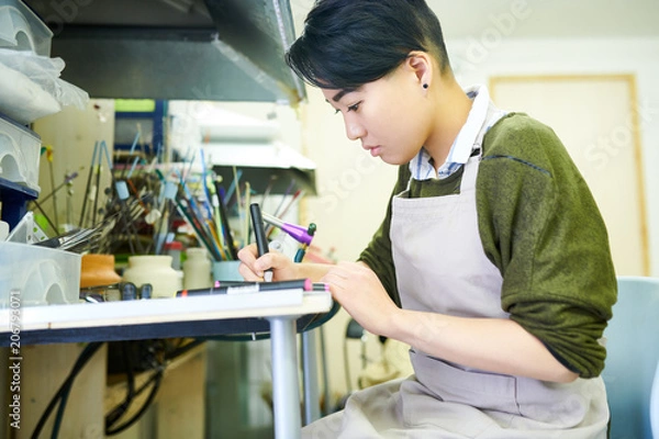 Obraz Side view portrait of young Asian woman drawing sketches in glassworking workshop, ready to create beautiful jewelry