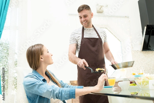 Obraz Cheerful excited handsome young man in apron holding cooking pan and putting food in plate while feeding girlfriend at home trying to surprise her
