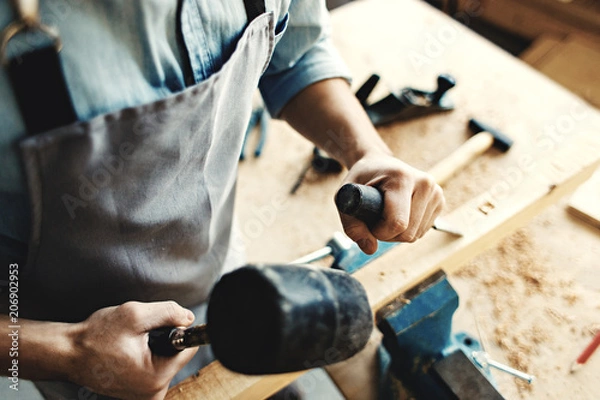 Obraz Hands of skillful woodworker using hammer and chisel to make mortise in wood plank, high angle view.