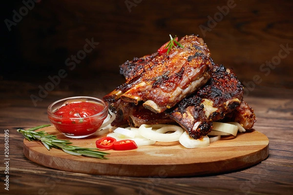 Obraz Fried ribs with rosemary, onion, sauce on a wooden round Board. Dark background. Place for text, copyspace