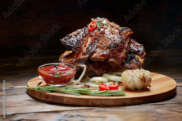 Fototapeta Fried ribs with rosemary, onion, sauce on a wooden round Board. Dark background. Place for text, copyspace