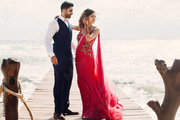 Obraz Wind blows bride's pink sari while she stands with Hindu groom on a wooden quay among foaming waves