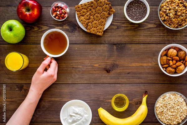 Fototapeta Set of products for healthy vegetarian breakfast. Fruits, oatmeal, yogurt, nuts, crispbreads, chia on dark wooden background top view space for text. Hand takes tea