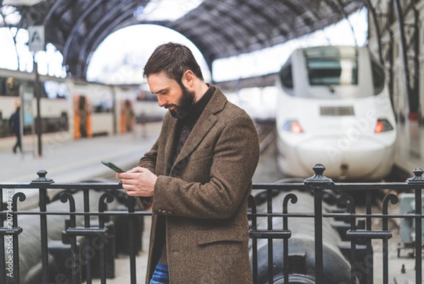 Fototapeta Attractive adult man using mobile application on his smartphone at train station.Business travel concept