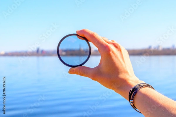 Fototapeta Female hand with a photo filter on the background of the lake and the city