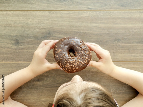 Obraz Child eating a donut on wooden background with copy space. Unhealthy eating concept, top view