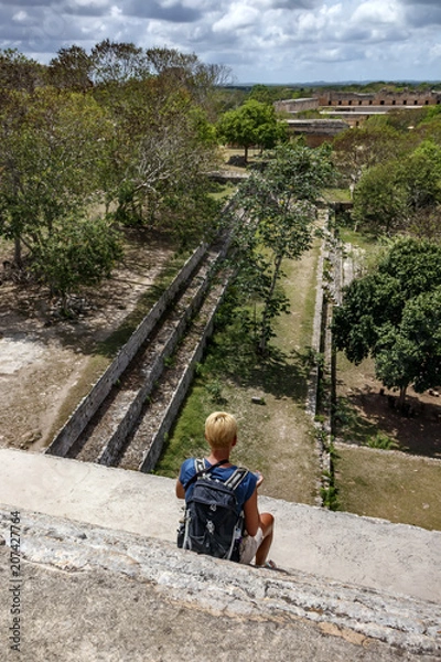Fototapeta Girl sitting on the steps of the pyramid and looking at the ancient city of Maya Uxmal. Mexico