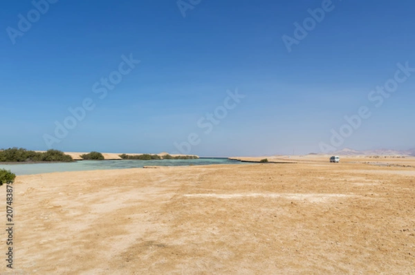 Fototapeta Desert landscape, with a sea estuary, and mangrove trees growing on it, a minibus in the background is driving along a dirt road, against a blue sky and mountains, Ras Mohammed, Egypt.