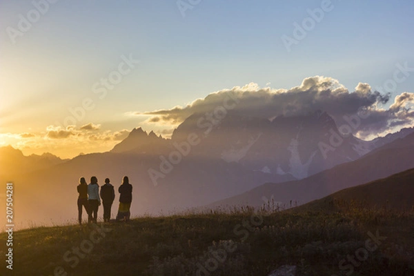 Obraz group of people with hands up standing on grass in sunset mountains