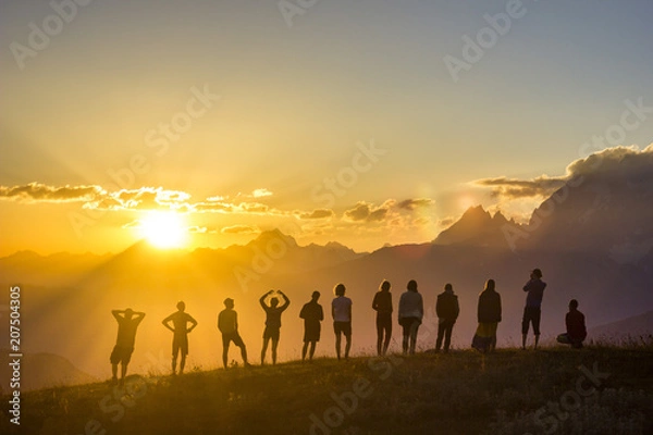 Obraz group of people with hands up standing on grass in sunset mountains