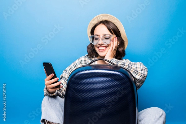 Obraz Young pretty woman in sunglasses and straw hat with a suitcase looks into the phone isolated on blue background. Flight traveling.