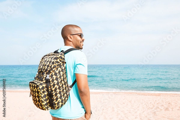 Fototapeta Portrait of attractive young African-American hipster wearing t-shirt and sunglasses walking at summer day on the beach with backpack