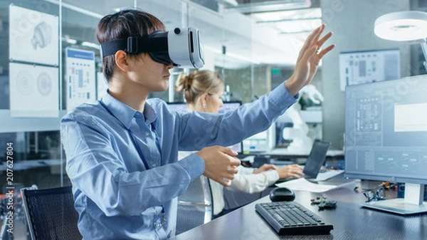 Fototapeta Computer Science Engineer wearing Virtual Reality Headset Works with 3D Modeling, Makes Gestures. In the Background Engineering Bureau with Busy Corowrkers.