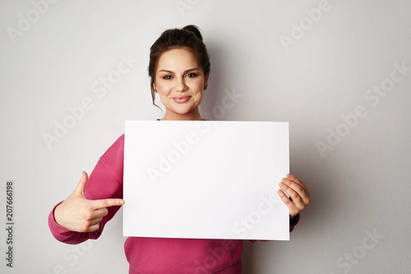 Fototapeta Pretty smiling woman in pink hoody holding empty blank board isolated on the gray background