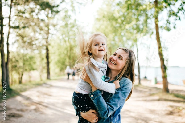 Obraz Happy smiling mother hugging her lovely little daughter outdoor. Lifestyle family. Adult cheerful female parent playing with her beautiful emotional child at nature in summer. Positive people faces.