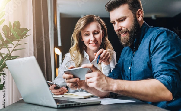 Fototapeta Business meeting. Teamwork. Smiling businesswoman and businessman sitting at table in front of laptop. Man showing woman information on smartphone screen. Online marketing, e-learning, distance work.
