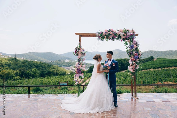 Fototapeta Newlyweds in love look at each other and enjoy the wedding day. They stand on an arch of pink, white and blue flowers.