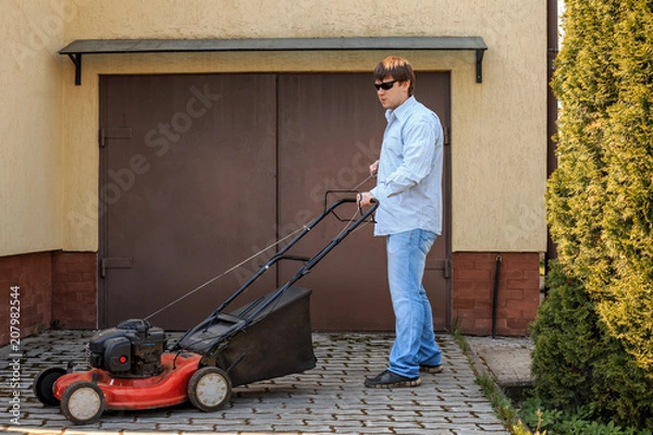Fototapeta Young man in sunglasses starts a lawn mower opposite the garage in the summer.
