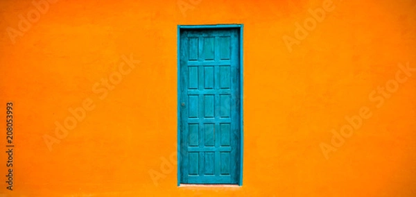 Fototapeta Vivid bright orange colour facade with blue-green closed door in the center of large empty orange wide wall texture background space in panorama format.