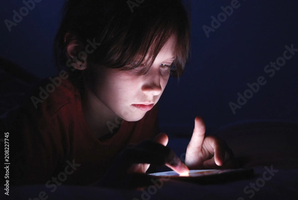 Fototapeta Caucasian girl lying in bed playing tablet in social internet in dark blue light under blacket. Childhood, communication and technology concept