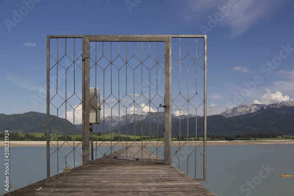 Fototapeta Looking through a locked lattice door onto a wooden boat dock across the dry Forgensee in the direction of the Alps