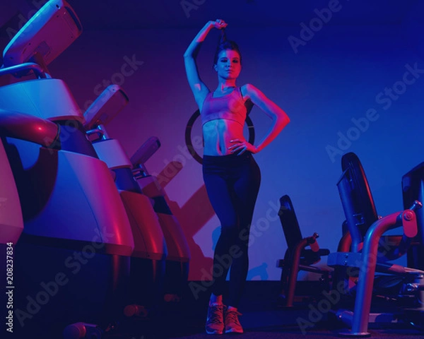 Fototapeta Waist up portrait of young attractive brunette woman dressed in sports clothes illuminated by faint pink light. Gorgeous confident slim female fitness model wearing top and leggings posing at gym.