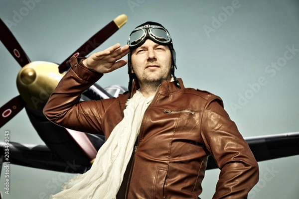 Obraz Portrait of a vintage pilot with leather cap, scarf and aviator glasses salutes in front of a historic airplane - Portrait of a man in historical pilot clothing