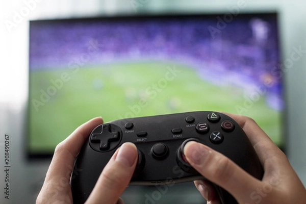 Fototapeta Female hands hold the joystick on the background of the TV, a game of football, close-up, a girl a gamer