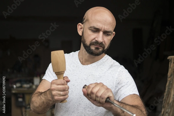 Fototapeta white bald carpenter with beard in white t-shirt works as a chisel and a mallet