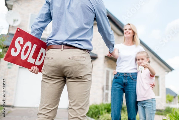 Fototapeta cropped shot of realtor with sold sign shaking hand of young woman with daughter in front of new house
