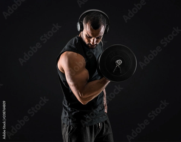 Obraz Strong handsome sport man making weightlifting, listening music and exercising the biceps on isolated black background