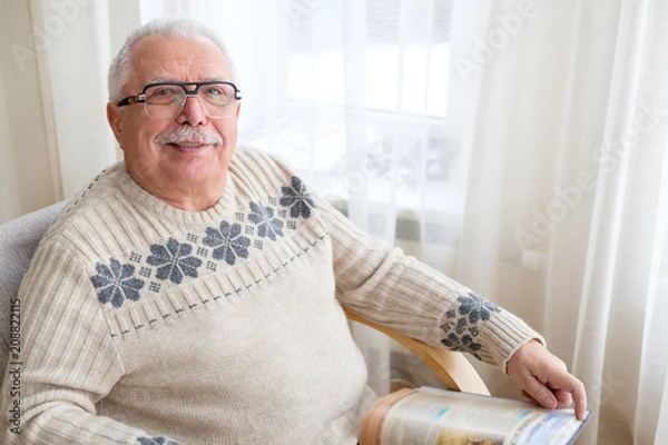 Fototapeta Senior man 75-75 years old sitting at home and reading book in armchair. Old age, lifestyle and people concept