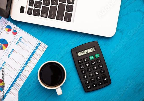 Fototapeta Investment, finance and work concept with charts and graphs and laptop, coffee, calculator on blue board flat lay