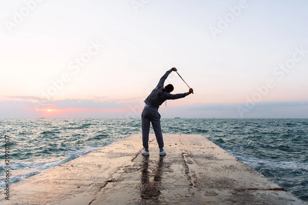 Fototapeta Full length photo of sportsman practicing exercises, stretching with skipping rope, standing on pier, near the sea, wearing sport clothes. Back view. Outdoors.