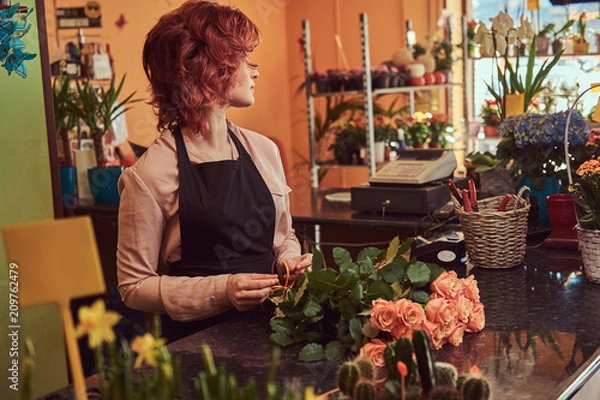 Fototapeta Charming redhead female florist wearing uniform making a beautiful flower composition while standing at the counter of flower shop.