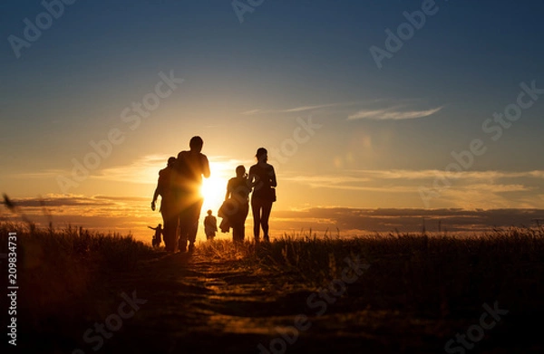 Fototapeta A group of people walking in a track. They go against the background of the orange sun, their contours and silhouettes are visible.
