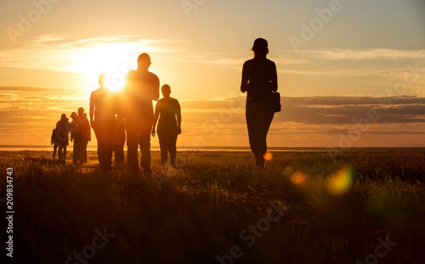 Fototapeta Grupa ludzi idących ścieżką. Idą na tle pomarańczowego słońca, widoczne są ich kontury i sylwetki.