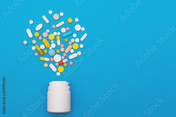 Obraz Assorted medical pills and capsules scattered from a bottle on a blue background. Empty place for text. Background.