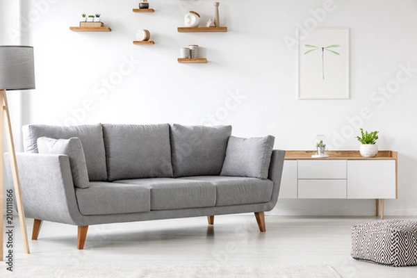 Fototapeta Simple, gray sofa standing next to a white cupboard in living room interior with decorations on wooden shelves. Real photo