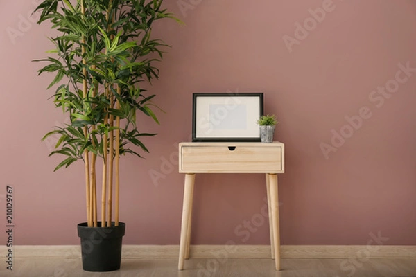 Fototapeta Wooden table with frame and green plant near color wall