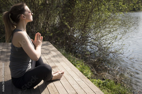 Fototapeta Young girl practices yoga on the shore of the lake, the concept of enjoying privacy and concentration, sunlight