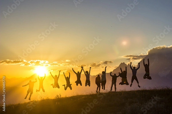 Obraz group of people with hands up jumping on grass in sunset mountains