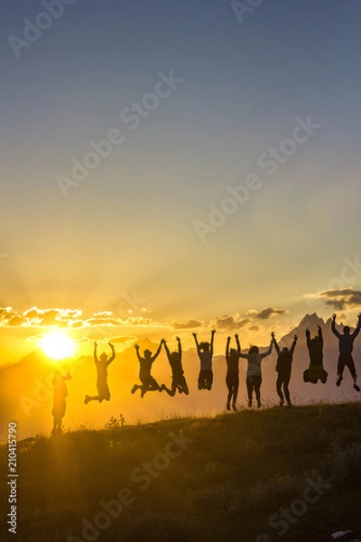 Obraz group of people with hands up jumping on grass in sunset mountains